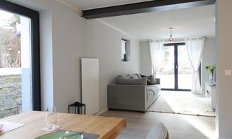 A bright and modern living atmosphere with a gray sofa and large windows. The space is open and inviting, with a dining table in the foreground.