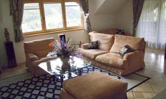 A cozy living room with beige sofas and a glass table. Large windows let in a lot of light and offer a view of nature.