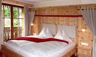 A cozy bedroom with a large wooden bed and two pillows. The walls are clad in wood, and there are large windows that allow plenty of light into the room.