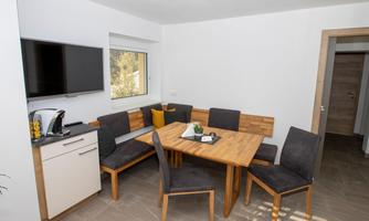 A modern dining area with a wooden table and black chairs. On the wall, there is a television and a window looking outside.