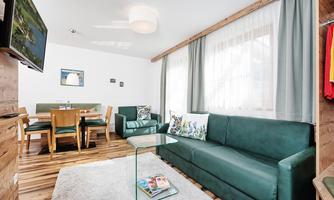 A modern living room with a green sofa and a glass coffee table. Inside, there is a dining area with wooden tables and chairs, as well as large windows that let in plenty of daylight.