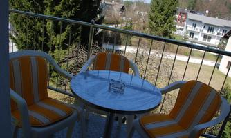 A cozy balcony with a round table and two chairs in orange and white. In the background, trees and other buildings can be seen.