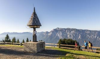 A group of hikers is walking at a viewpoint with a small pavilion. In the background, majestic mountains and a clear sky can be seen.