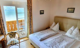 A cozy bedroom with two beds and a balcony overlooking a lake. The decor is bright and inviting, with a charming selection of chairs and decorations.