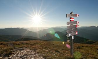 A signpost on a mountain with a shining sun in the background. The landscape is hilly and offers a vast view.