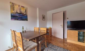 A cozy dining area with a wooden table and chairs. A colorful art print hangs on the wall and a television is in the corner.