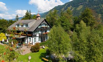 A beautiful building in alpine style, surrounded by trees and mountains. The sky is blue and the landscape looks inviting.