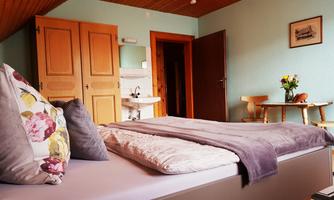 A cozy bedroom with a large bed and colorful pillows. In the background, there is a small table and a water trough.