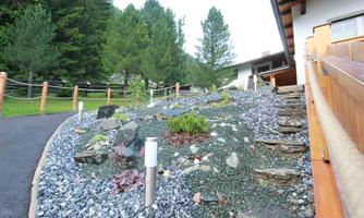 A beautifully designed garden with a gravel-covered slope and small plants. In the background, tall trees can be seen, and the path leads to the terrace of the house.