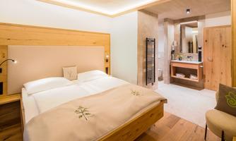 A modern bedroom with a large double bed and wood-paneled walls. In the background, there is a bathroom with stylish touches.