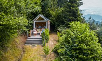 A small wooden structure among trees with a terrace. Two people are sitting relaxed in front of it, enjoying nature.