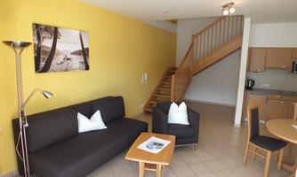 A cozy living room with a black couch and a wooden chair. The walls are painted yellow and a staircase leads upstairs.