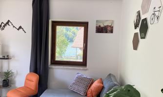 A cozy living room with a gray sofa and orange cushions. A window lets in a lot of daylight and there are decorative pictures on the wall.