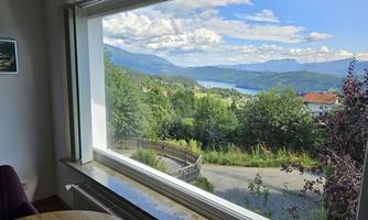 A view from a window of a picturesque landscape with mountains and a lake. The sky is blue with some clouds, and many trees and houses are visible.