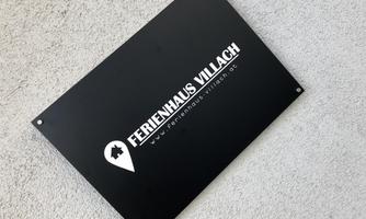 A black sign with the inscription "HOLIDAY HOUSE VILLACH" and a location marker. The sign is modern and easy to read.