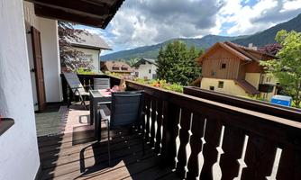 A balcony with a table and chairs, surrounded by houses and trees. In the background, mountains and a beautiful sky with clouds can be seen.