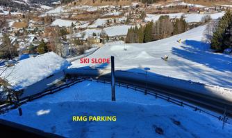 A snowy landscape with a ski trail and a parking lot. In the background, gentle hills and trees are visible.