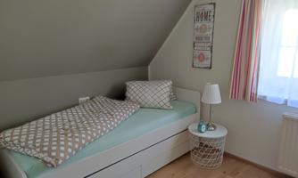 A bright bedroom with a single bed, decorative cushions, and a bedside table. A window with curtains lets in plenty of light.