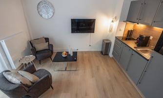 A modern living room with a television and an open kitchen. The room has light wooden flooring and a cozy atmosphere.
