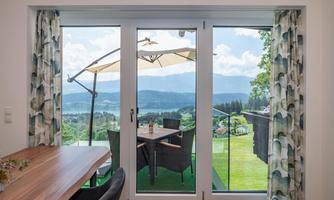 A modern balcony overlooking a green landscape and mountain ranges. A table with chairs and a sunshade invites relaxation.