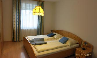 A cozy bedroom with a double bed and blue pillows. Large windows with curtains let in plenty of light.