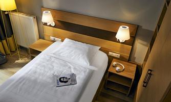 A modern bedroom with a single bed and wooden paneling. Next to the bed is a nightstand with a lamp and a book.
