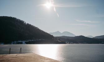 A quiet lake surrounded by mountains and trees. The sun shines in the clear sky and reflects in the water.