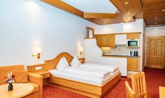 A cozy room with a double bed and a small kitchen area. The furniture is made of wood and the walls are painted light.