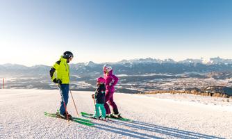 A family is standing on the ski slope in the snow. In the background, the mountains and the clear sky can be seen.