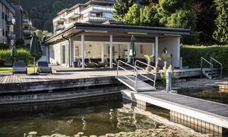 A modern building by the water with a wooden deck and relaxing lounge chairs. Access is via a walkway, surrounded by green plants.