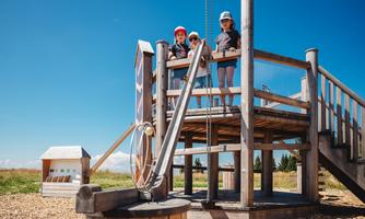 A playground with a large wooden play tower. Three children are standing on it, enjoying a clear blue sky in the background.