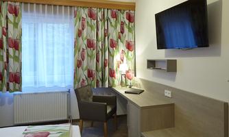 A cozy hotel room with colorful curtains and a television on the wall. There is a desk and a chair next to a window with natural light.