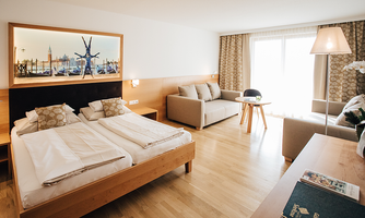 A modern hotel room with two single beds and a cozy seating area. Large windows provide plenty of daylight and a pleasant atmosphere.