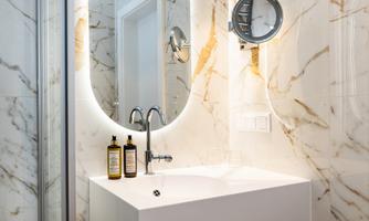 A modern bathroom with marbled walls and an elegant sink. In the background, there is a lit mirror and some bottles of toiletries visible.