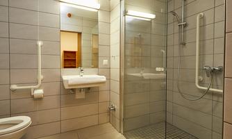 A modern bathroom with a shower, a sink, and a toilet. The walls are clad with light tiles and there are handrails for additional safety.