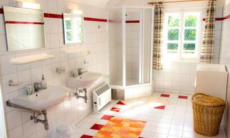 A bright bathroom with two sinks and a shower. Red and white tiles, along with a braided rug, add color accents to the space.