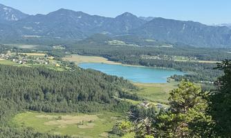 A picturesque landscape with a clear lake, surrounded by green forests and mountains. The sky is blue and cloudless.