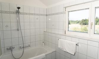 A modern bathroom with white tiles. There is a bathtub, a shower, and a window with a view of the greenery.