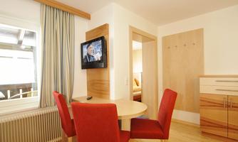 A cozy living area with a round table and red chairs. There is a TV on the wall and the room has light wooden flooring.