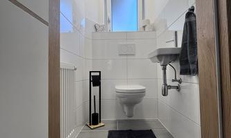 A modern bathroom with white tiles and a bright window. In the foreground, there is a black rug and a towel holder.