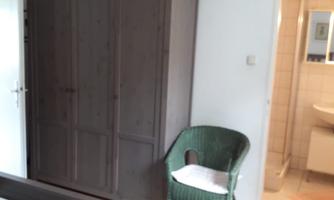 A simple bedroom with a wooden wardrobe and a green chair. In the background, there is a bathroom and two beds with bedding.