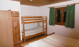 A simple room with a bunk bed and a double bed. There are windows with green curtains and a wooden wardrobe.