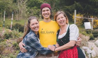 Three people are smiling in a green garden landscape. They are wearing colorful clothing and seem happy together.