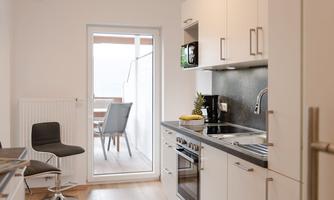 A modern kitchen with light furniture and a window leading to a balcony. Some fresh fruits are on the countertop.