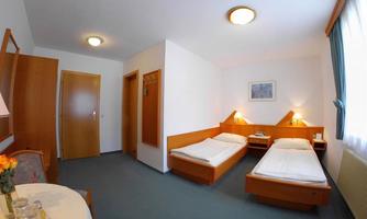 A simple guest room with two single beds and a small table. The walls are light, and the room has a window with curtains.