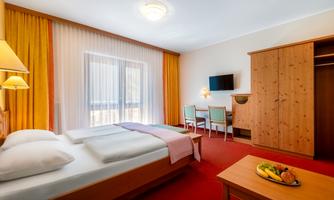 A bright hotel room with two beds and a wooden table. The room features large windows and a cozy atmosphere.