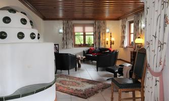 A cozy living room with a bright wooden ceiling and comfortable black sofas. A decorative tiled stove and windows with curtains can also be seen.