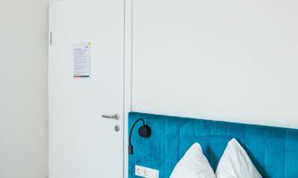 A modern bedroom with a blue headboard and two white pillows. The walls are light and there is a door in the background.
