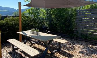 A shady picnic area with a table and benches under a sunshade. Surrounded by green plants and wood chips.