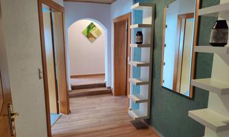 A bright hallway with wooden flooring and white walls. Shelves and a mirror stand against the wall, while a staircase leads to the next room.